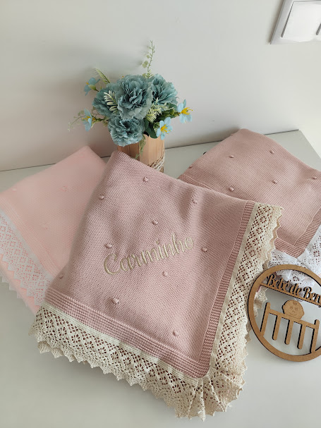 Três panos de cozinha rosa e bege com renda e bordado ao lado de flores azuis e suporte de madeira sobre mesa branca.