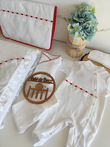 Conjunto de roupa de bebé branca com detalhes em renda branca e fita vermelha, com vaso decorativo de flores azuis ao lado