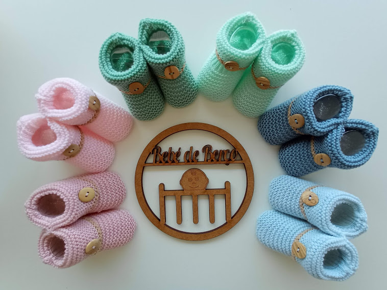 Vários sapatinhos de bebé em crochet de cores pastel dispostos em círculo em torno de um ornamento de madeira com texto.