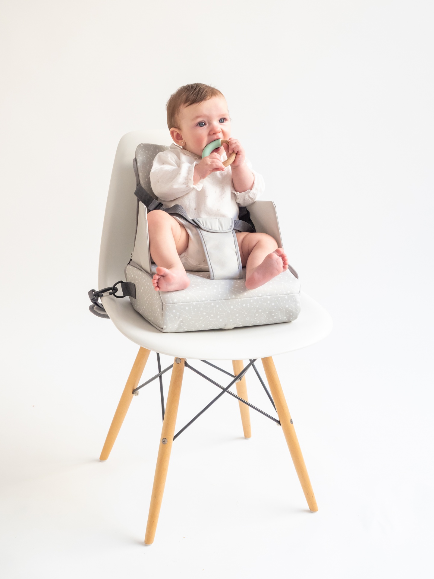 Bebé em assento elevatório cinzento com padrão de pontos numa cadeira branca de pernas de madeira