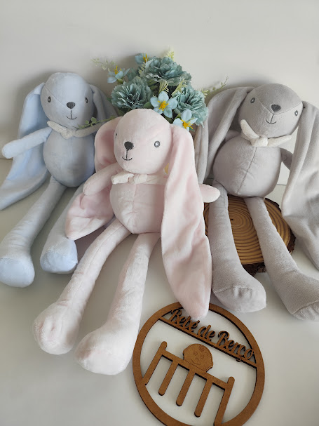 Três coelhos de peluche coloridos com orelhas longas, arranjo floral e placa de madeira gravada