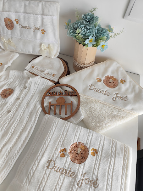 Conjunto de produtos de bebé em tecido cru com bordados castanhos de leão e texto Duarte José, junto a vaso de flores azuis.