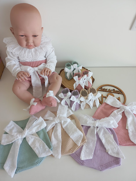 Boneca de bebé com roupa branca e rosa e roupas de malha coloridas com fitas grandes brancas