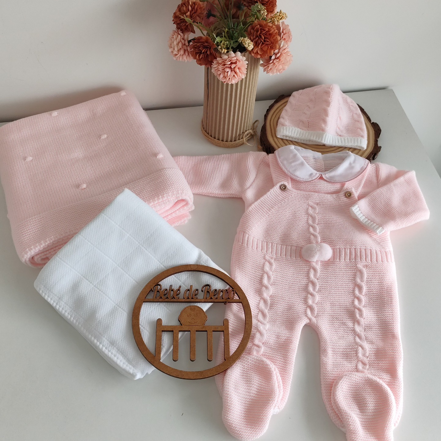 Conjunto de roupa de bebé rosa de malha com manta, gorro e fralda numa mesa branca com vaso de flores