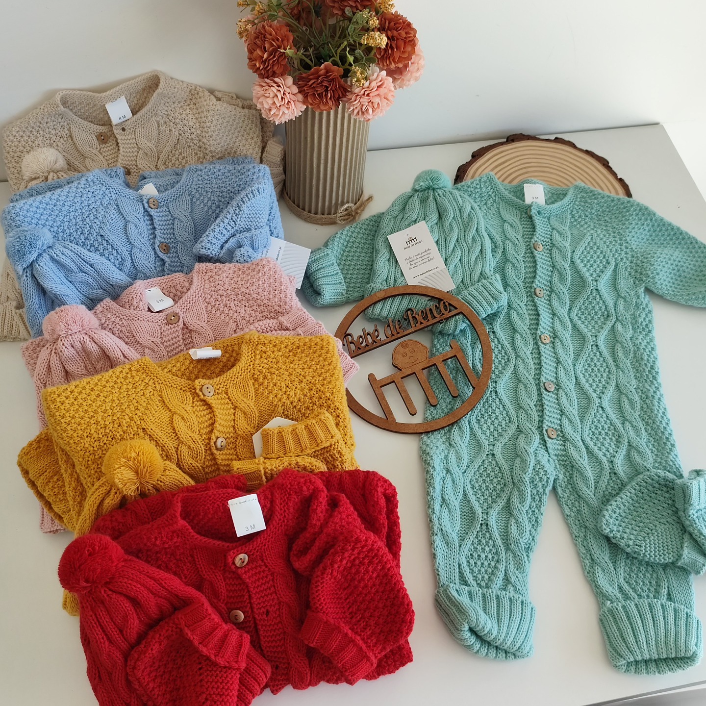 Roupas de bebê em malha tricotada de várias cores sobre fundo branco com vaso de flores e placa de madeira