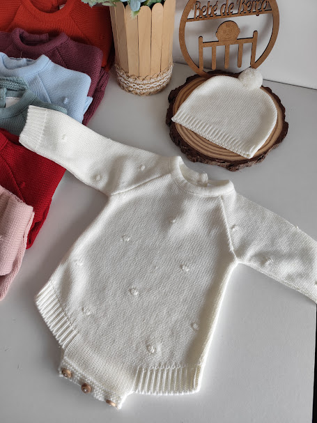 Conjunto de bebé branco em malha com casaco e gorro, várias roupas coloridas ao lado e decoração em madeira