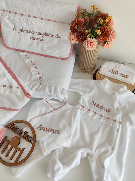 Conjunto de roupa de bebé branca com detalhes cor-de-rosa e flores num vaso à direita