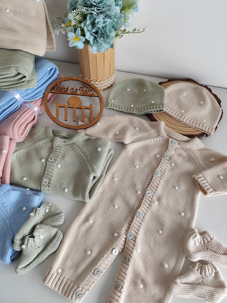 Roupas de bebé em malha creme, verde e azul com bolinhas, placa de madeira e vaso de flores