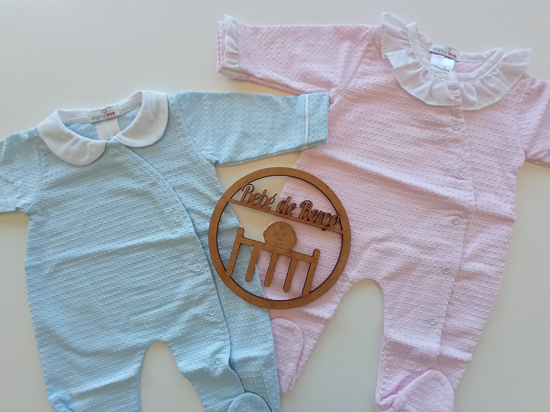 Dois macacões para bebé, um azul e outro rosa, com gola branca, e objeto redondo de madeira entre os macacões.