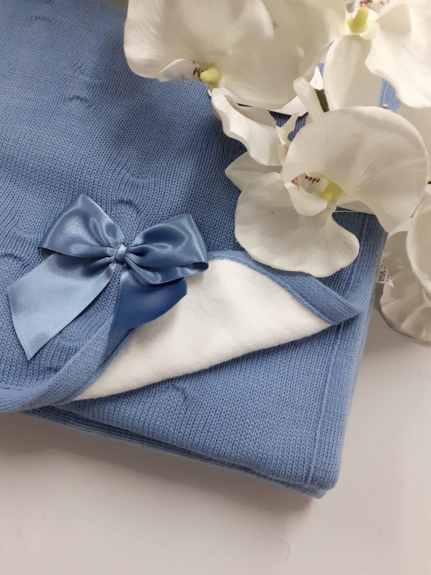 Manta azul clara de malha com laço azul de cetim ao lado de orquídeas brancas