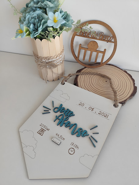 Placa decorativa com informações de nascimento, vaso com flores artificiais e enfeite de madeira em mesa