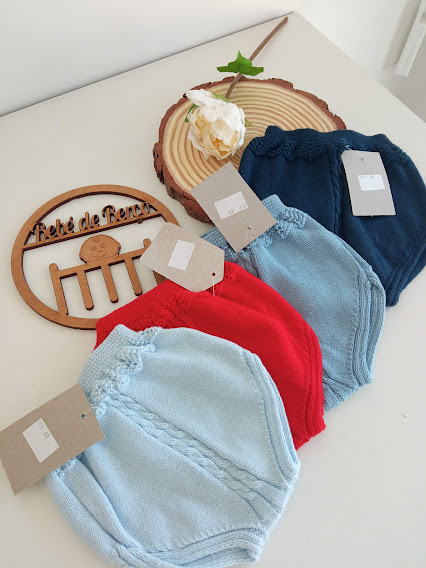 Peças de roupa de malha de bebé em várias cores com etiquetas e placa decorativa 'Bebé de Renas' numa mesa.