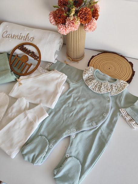 Conjunto de roupa para bebé com macacão verde água, camisola branca, toalha branca bordada e vaso com flores secas.