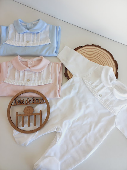 Três macacões de bebé nas cores azul, rosa e branco com aplicações brancas e botões, sobre superfície branca e base de madeira com placa decorativa