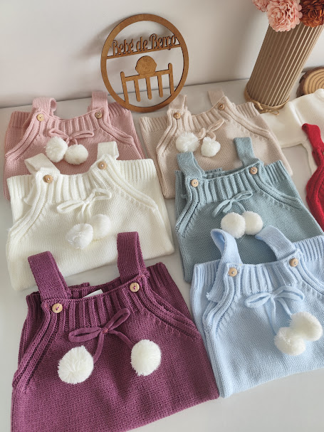 vestidos de malha para bebé em várias cores com pompons e botões de madeira sobre superfície branca