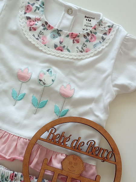 T-shirt de bebé branca com flores bordadas, gola floral e folho rosa, etiqueta de algodão 1M, com placa de madeira 'Bebé de Bença' em cima.