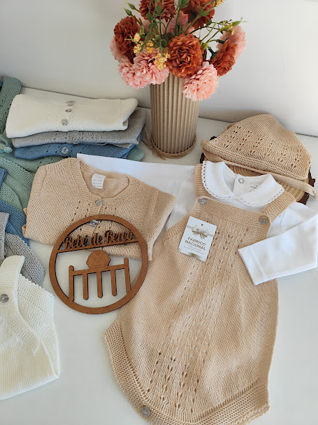 Roupas de bebé de malha em tons beges e azuis com vaso de flores secas ao fundo