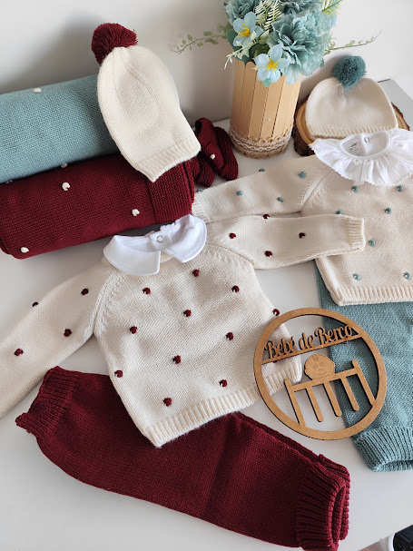 Conjunto de roupa de bebé com malhas creme, vermelhas e azuis e flores artificiais num vaso.