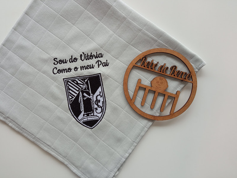 Toalha branca com bordado preto e brasão, ao lado de decoração redonda de madeira com texto.