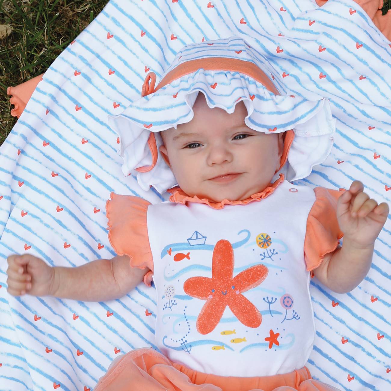 bebé com conjunto laranja e chapéu branco com riscas azuis e corações cor-de-laranja