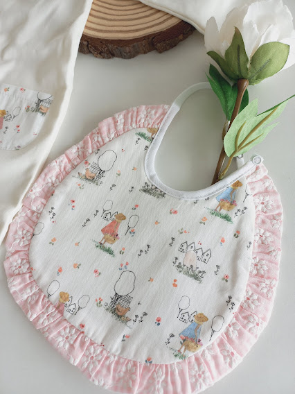 Babete de bebé branco com padrão infantil e borda rosa colocada em mesa branca com flores.