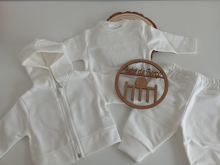 Conjunto de roupa branco para bebé com sweat, camisola e calças exposto sobre superfície clara com decoração em madeira.