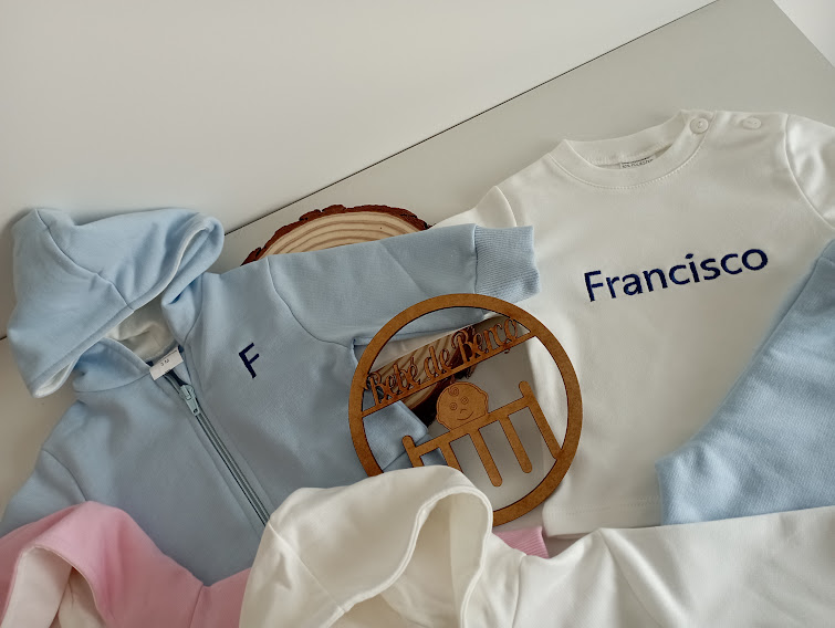 Roupas de bebé em azul claro, branco e rosa, incluindo camisola com nome 'Francisco' e objeto decorativo de madeira circular
