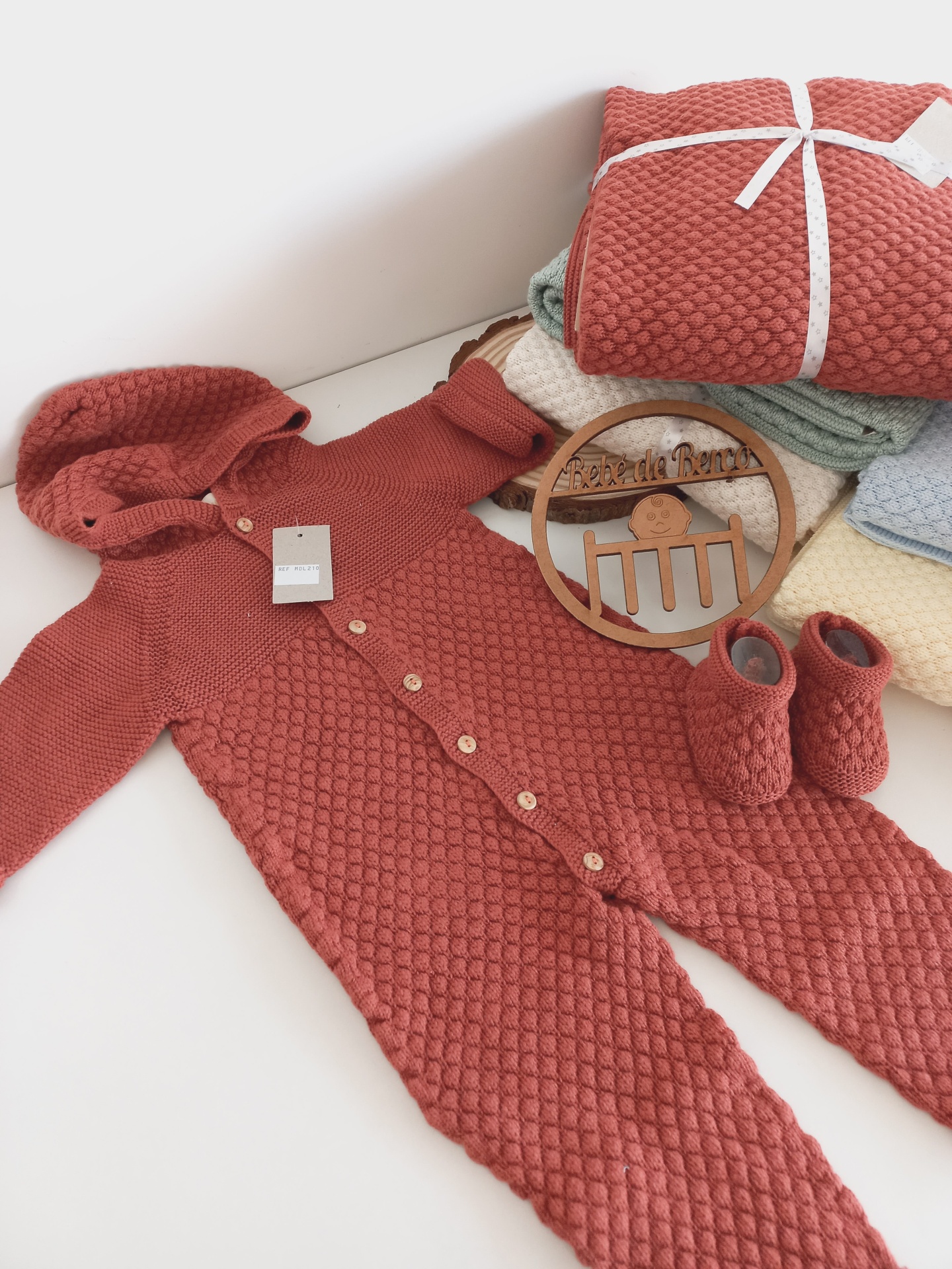 Fato de bebé vermelho em malha texturizada com capuz e sapatinhos, com toalhas coloridas e fato enrolado com fita branca ao fundo.