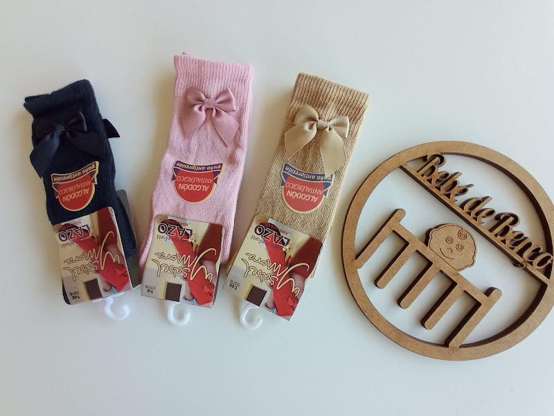 Três pares de meias de bebé azul escuro, rosa e bege com laço e etiquetas, e enfeite de madeira 'Bebe a Bordo'