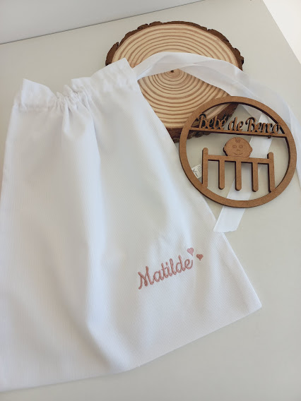 Saco branco com nome Matilde bordado ao lado de medalhão de madeira com texto e desenho de berço