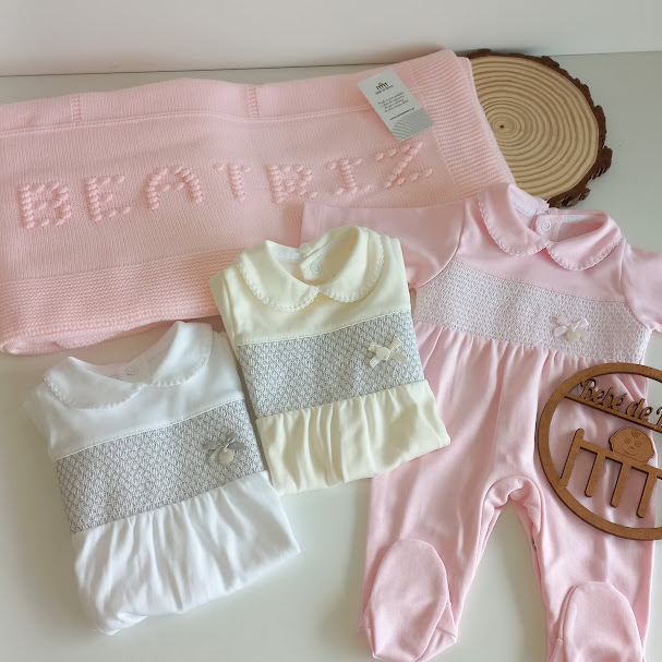 Manta rosa com o nome BEATRIZ e três roupas de bebé sobre superfície branca com placa de madeira