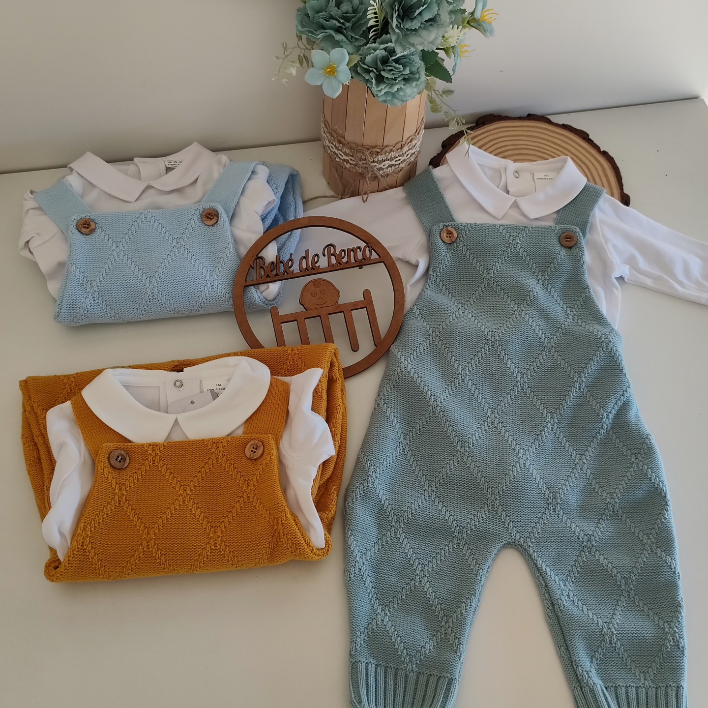 Conjuntos de roupa de bebé com jardineiras de malha branco, azul claro e mostarda, e decoração de flores em vaso