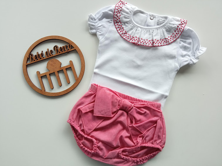 Conjunto de roupa de bebé em branco e cor-de-rosa com body e cueiros, junto a placa redonda de madeira com texto
