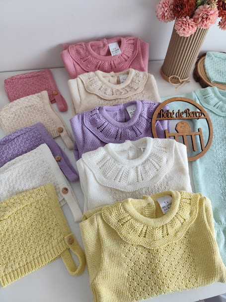 Conjunto de malhas para bebé em várias cores pastel com gola trabalhada e botões de madeira sobre uma mesa branca.