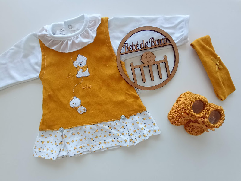 Conjunto de roupa de bebé com vestido amarelo mostarda, camisa branca, meias e sapatinhos amarelos, com placa de madeira Bebé de Pernas.