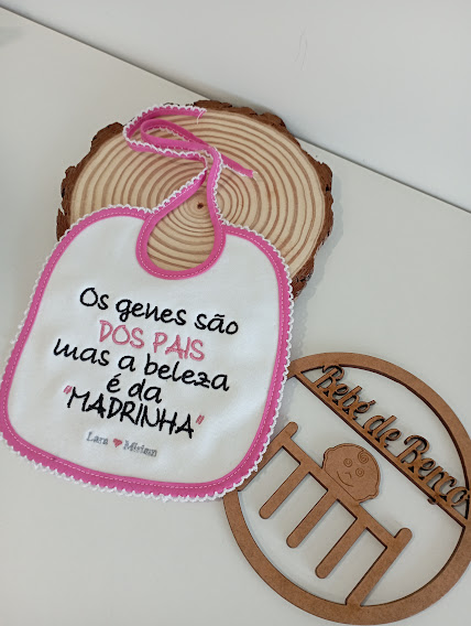 Babete branco com bordas cor de rosa e texto bordado, colocado sobre uma base redonda de madeira e adereço de madeira com texto