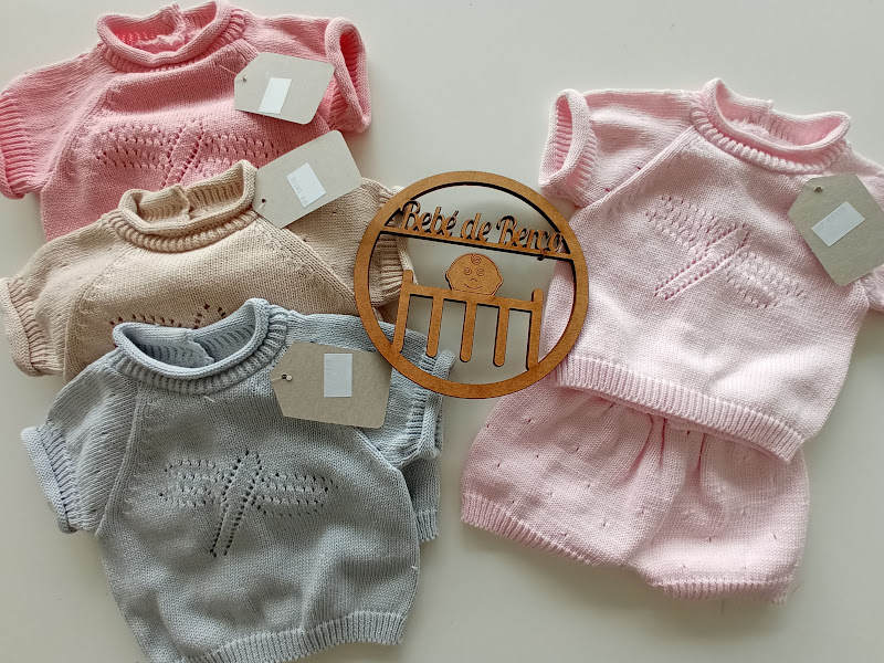 Quatro conjuntos de roupa de bebé em malha colorida com padrão rendado e etiquetas, sobre fundo branco com objeto decorativo de madeira