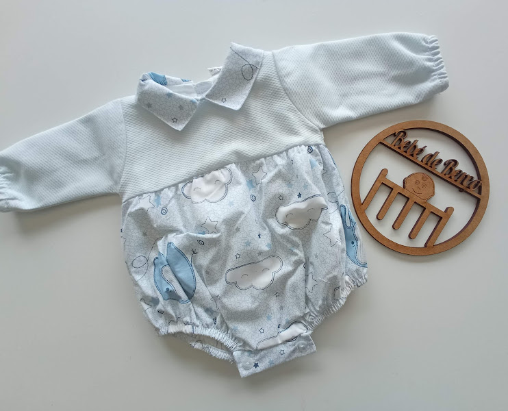 Body de bebé azul e branco com padrão de nuvens e aviões ao lado de objeto redondo de madeira