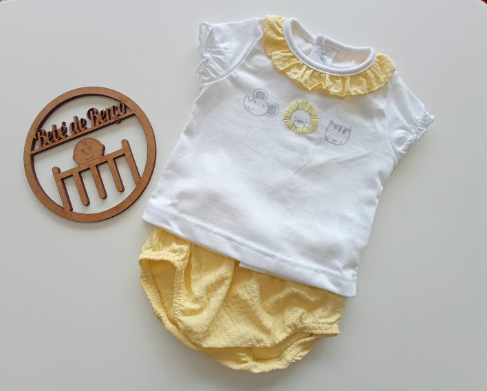 Conjunto de roupa de bebé branca e amarela com gola folhada e estampados, ao lado de objeto decorativo de madeira.