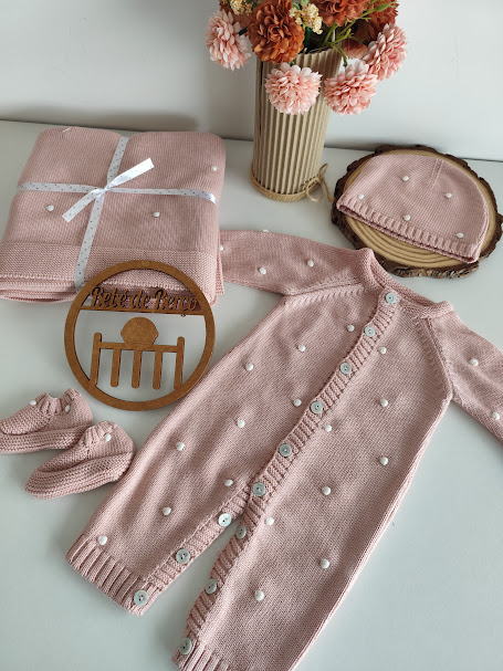 Conjunto de roupa de bebé rosa com bolinhas brancas, macacão, touca, pantufas e manta em superfície branca com vaso de flores