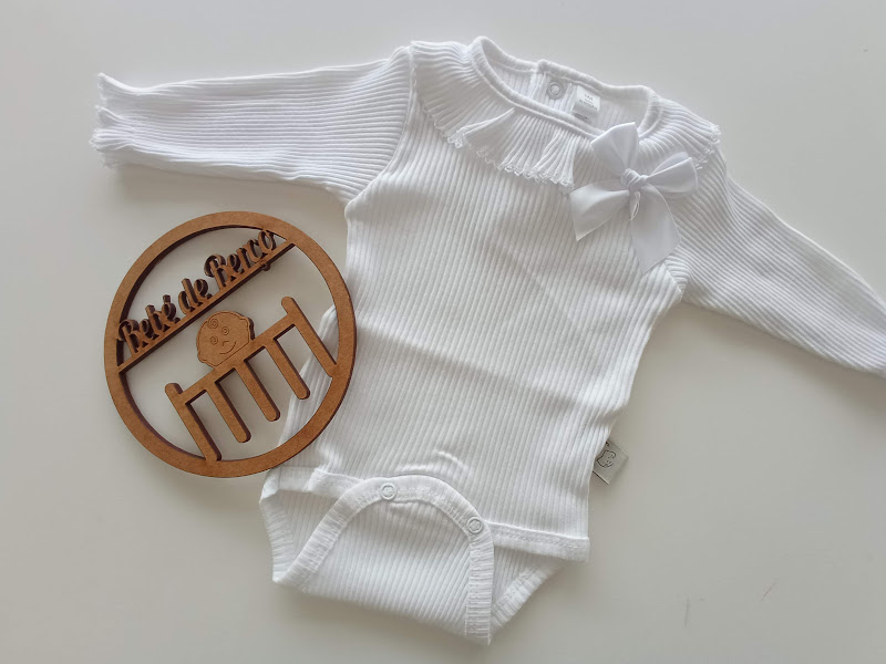 Body de bebé branco com manga comprida e laço ao lado de anel de madeira com texto