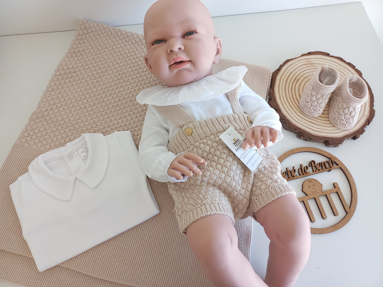 Boneco bebé vestido com roupa branca e bege, manta e decoração em madeira