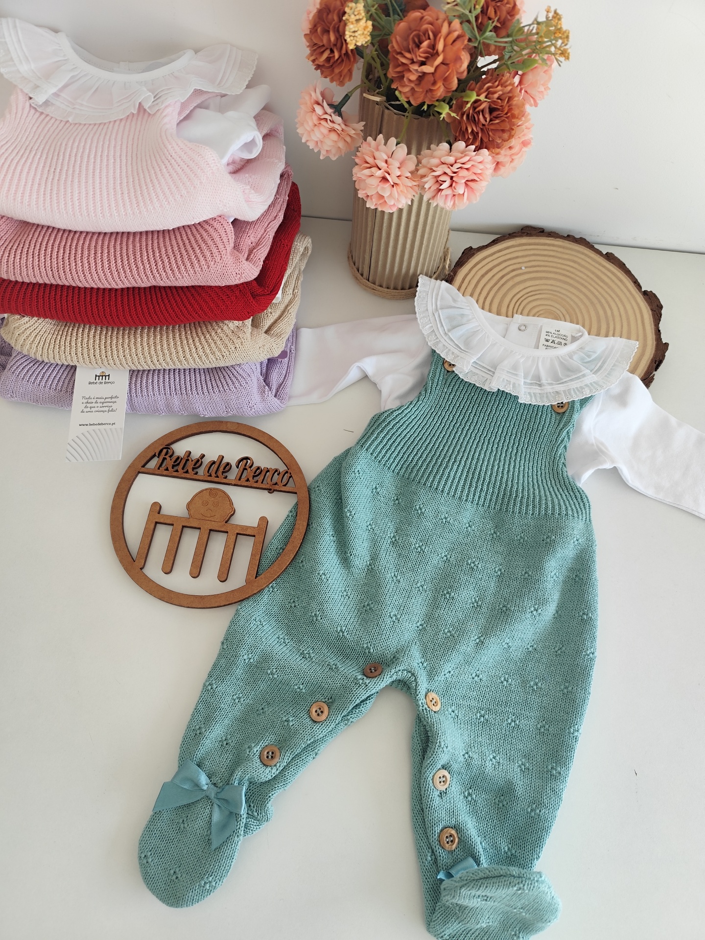 Conjunto de roupa de bebé com jardineira verde água e body branco junto a pilha de roupas coloridas e vaso com flores