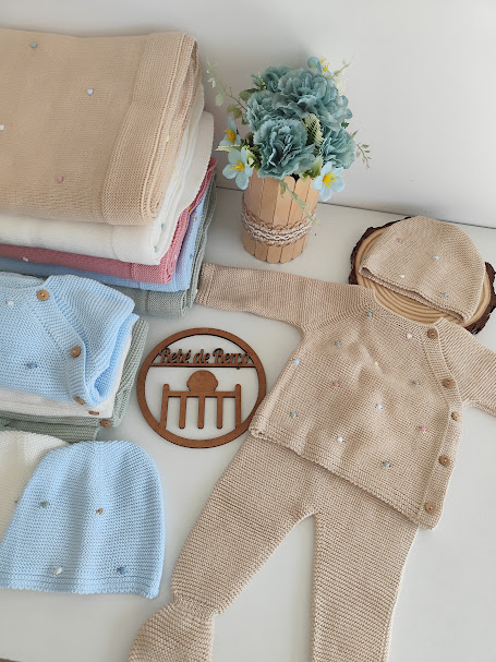 Conjunto de roupa de bebé em malha bege com botões e pontos coloridos, mantas empilhadas, chapéu, flores artificiais e placa de madeira com texto.
