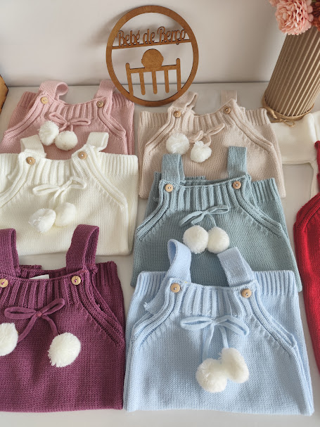 Peitorais de malha para bebé em cores variadas com botões e pompons, placa de madeira com texto ao fundo