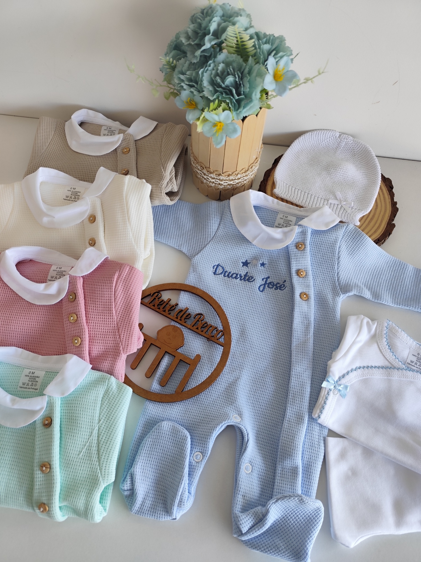 Roupa de bebé em várias cores com fio e bordados, disposta com flores e acessórios