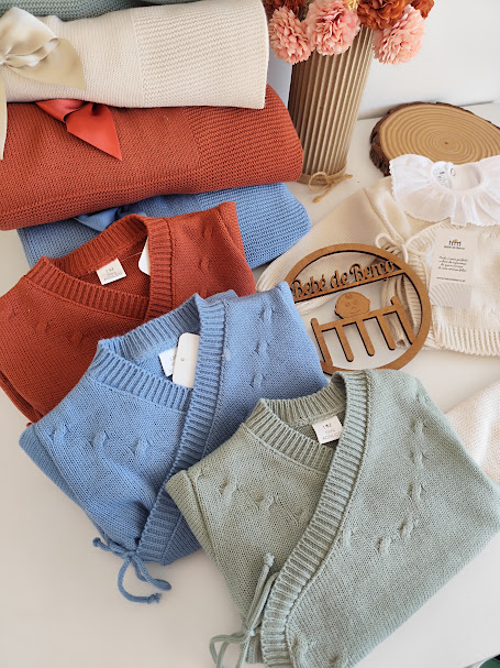 Peças de roupa infantil em malha de várias cores e textura tricotada dispostas sobre uma superfície branca com flores e um suporte de madeira decorativo