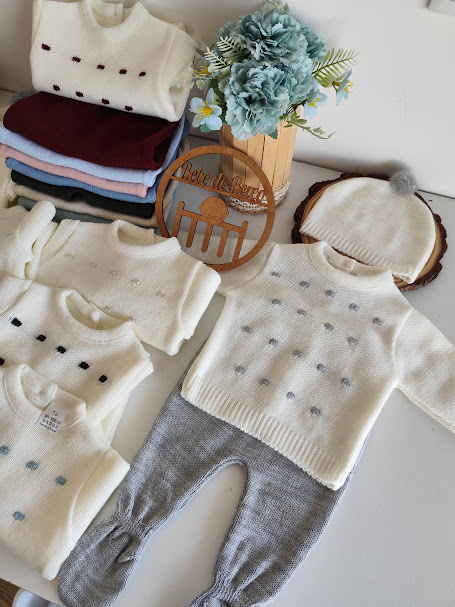 Roupas de bebé em tons neutros e pastéis com padrão de pequenos quadrados, gorro creme com pompom cinzento e flores azuis num vaso de madeira