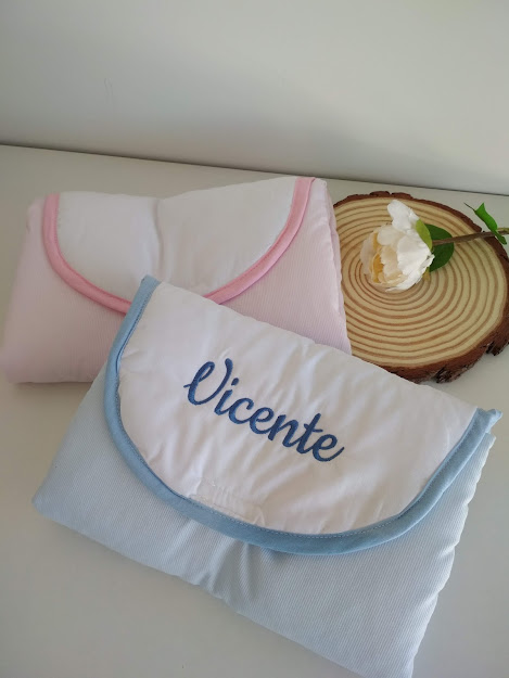 Duas toalhas para bebé, rosa e azul, com nome bordado 'Vicente' numa superfície branca com decoração de madeira e flor