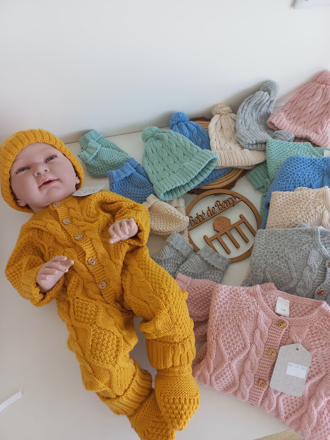 Bebé com macacão amarelo de malha e vários casacos e gorros de malha coloridos ao fundo